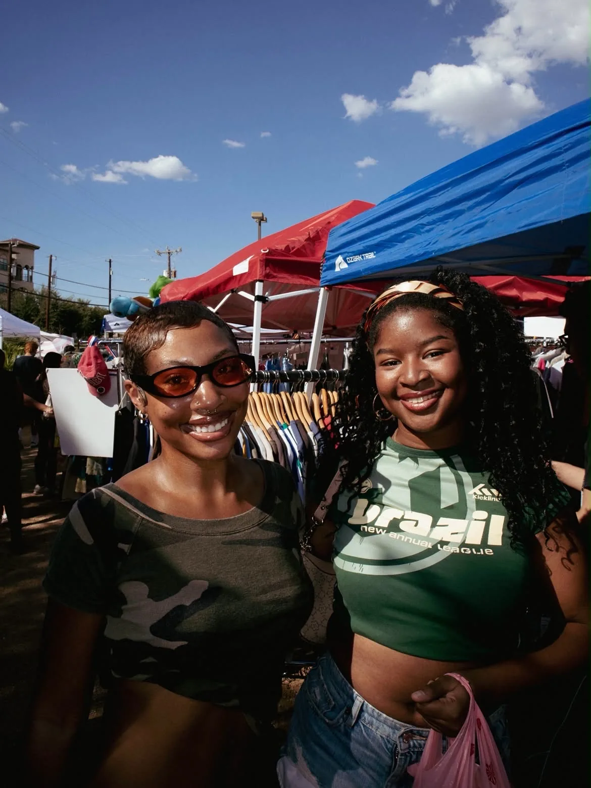 Two friends smiling in front of market canopies and clothing racks
